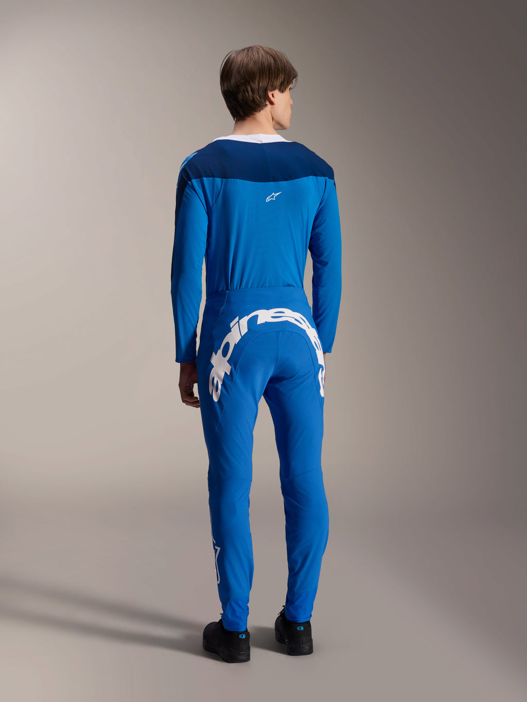 Modelo masculino visto de costas a usar a camisola de manga comprida Alpinestars A-Supra Melt azul, combinada com calças de performance azuis com a marca Alpinestars em branco na parte traseira, equipamento completo de corrida MTB