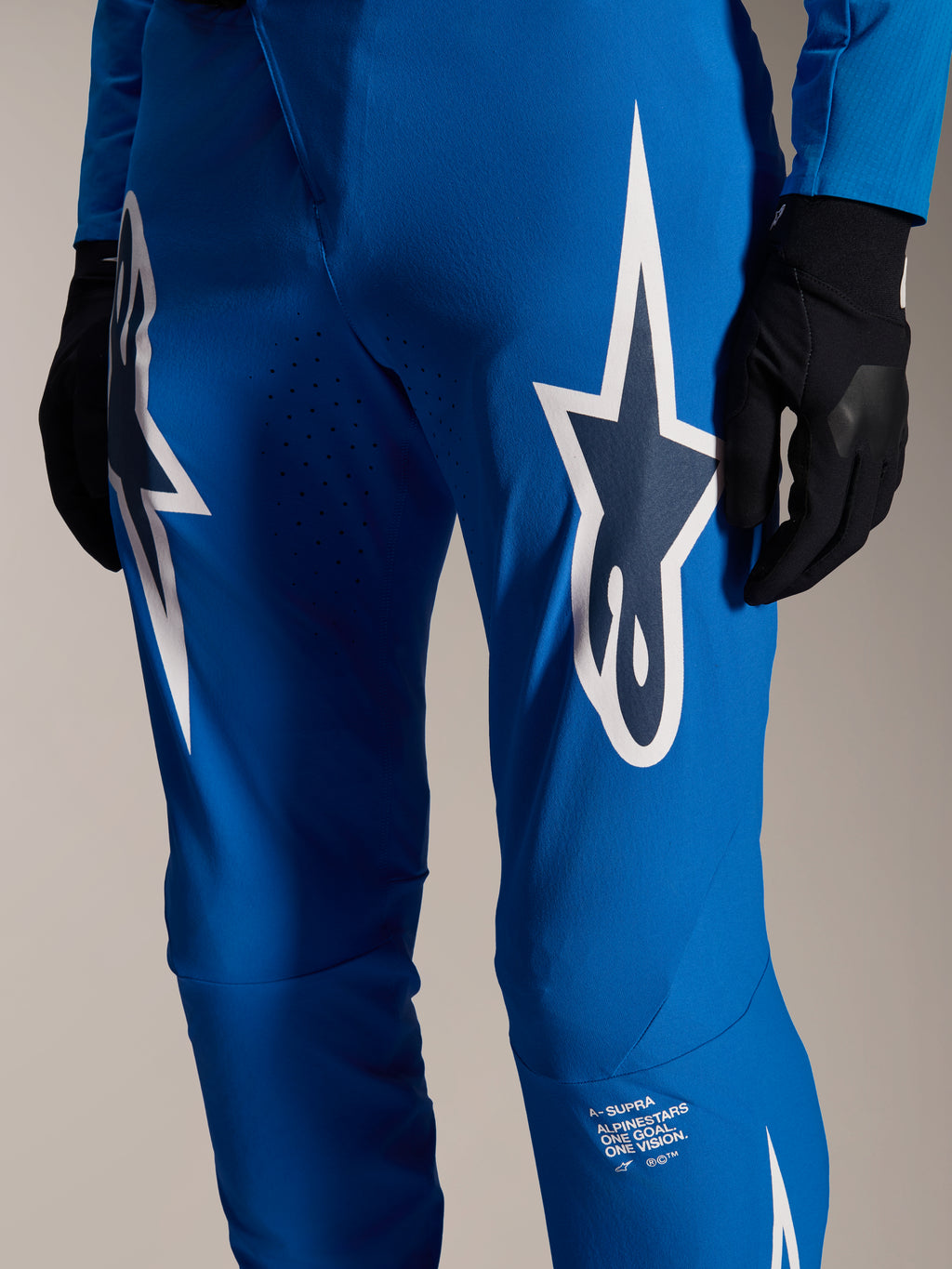 Calças Alpinestars A-Supra Melt, calças de ciclismo azuis, com grandes logótipos Astars brancos nas coxas, perfurações a laser para fluxo de ar e marca distintiva na parte inferior da perna, usadas com luvas pretas, equipamento de MTB de alta performance