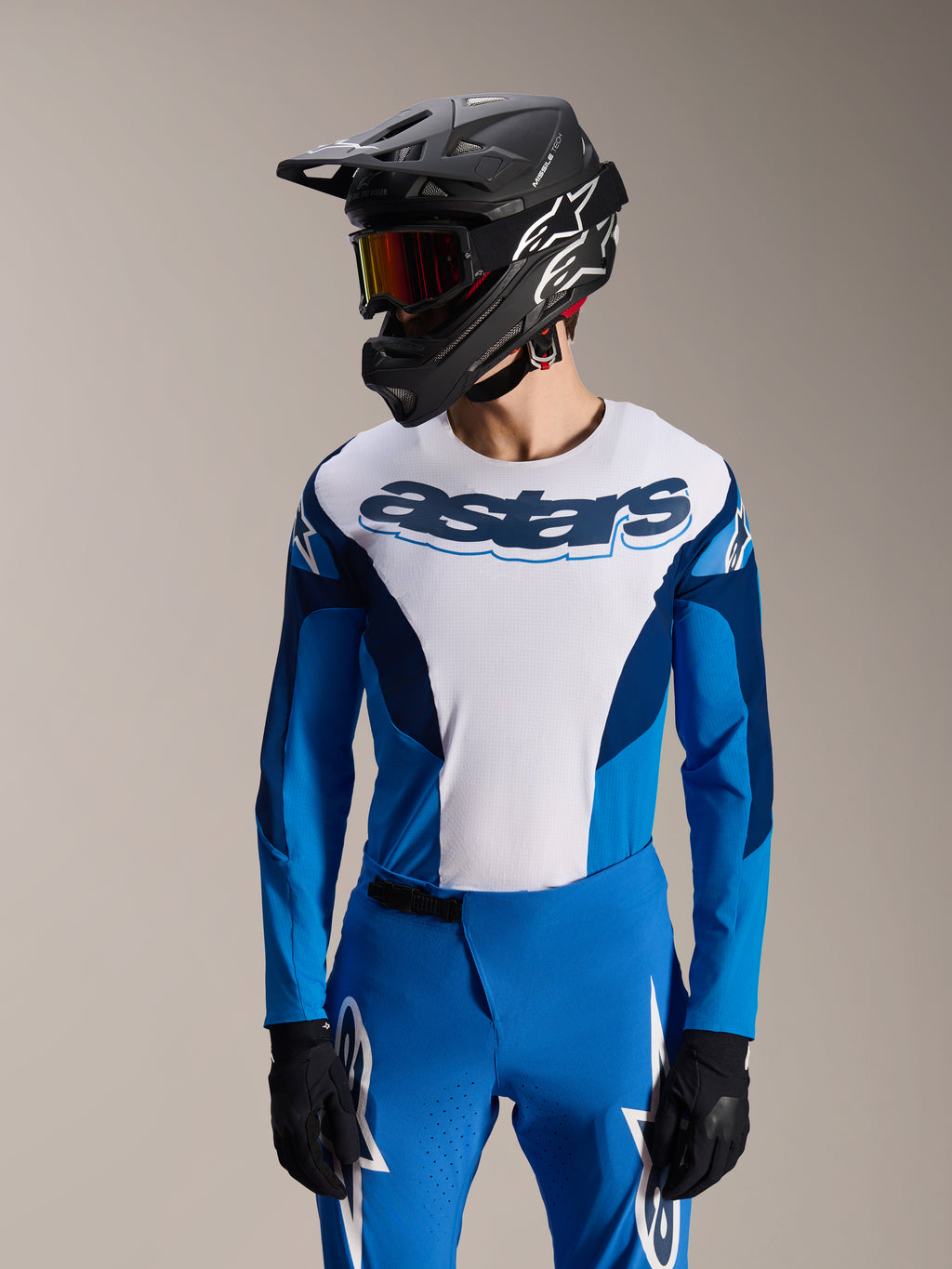 Modelo a usar camisola Alpinestars A-Supra Melt de manga comprida em azul e branco, com a marca nominativa 'astars' no peito, calças de BTT azuis a condizer, capacete integral preto e óculos de proteção, equipamento completo de corrida de BTT