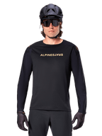 Jersey de Manga Longa Alpinestars A-Aria Polartec Switch, preta, logótipo Alpinestars dourado no peito, usada com capacete de BTT preto, óculos de sol escuros e luvas pretas, equipamento profissional para trilhos de mountain bike