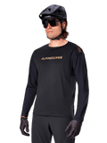 Jersey de manga comprida Alpinestars A-Aria Polartec Switch, preto com logótipo dourado, vestuário de BTT para ciclismo de montanha, usado com capacete preto e óculos de sol, com ventilação cortada a laser nas mangas e tecido Polartec com absorção de humidade para desempenho em todo-o-terreno