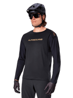 Jersey de manga comprida Alpinestars A-Aria Polartec Switch, preto com logótipo dourado, vestuário de BTT para ciclismo de montanha, usado com capacete preto e óculos de sol, com ventilação cortada a laser nas mangas e tecido Polartec com absorção de humidade para desempenho em todo-o-terreno