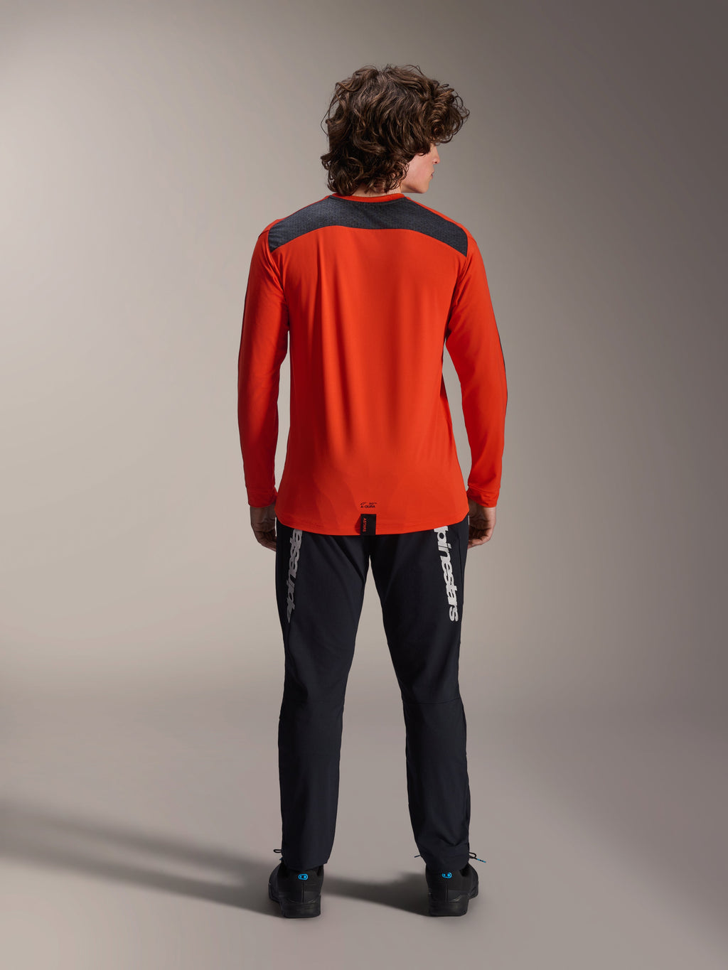 Alpinestars A-Dura Insert Astar Long Sleeve Jersey em Spicy Orange, vista de costas com painel de ombros em cinzento carvão, usada com calças Alpinestars pretas e sapatos pretos, fotografia profissional de estúdio para equipamento de performance de BTT