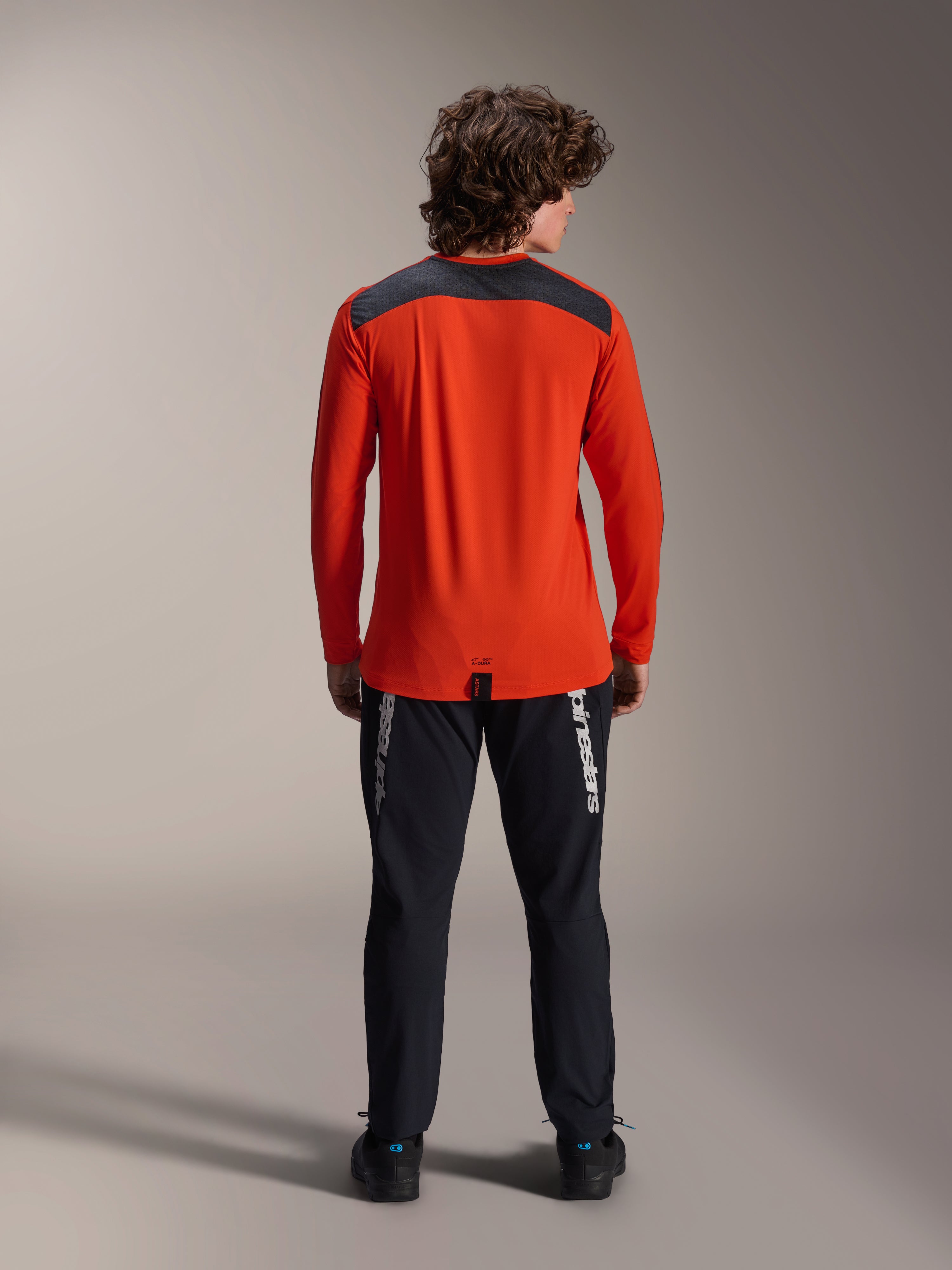Alpinestars A-Dura Insert Astar Long Sleeve Jersey em Spicy Orange, vista de costas com painel de ombros em cinzento carvão, usada com calças Alpinestars pretas e sapatos pretos, fotografia profissional de estúdio para equipamento de performance de BTT