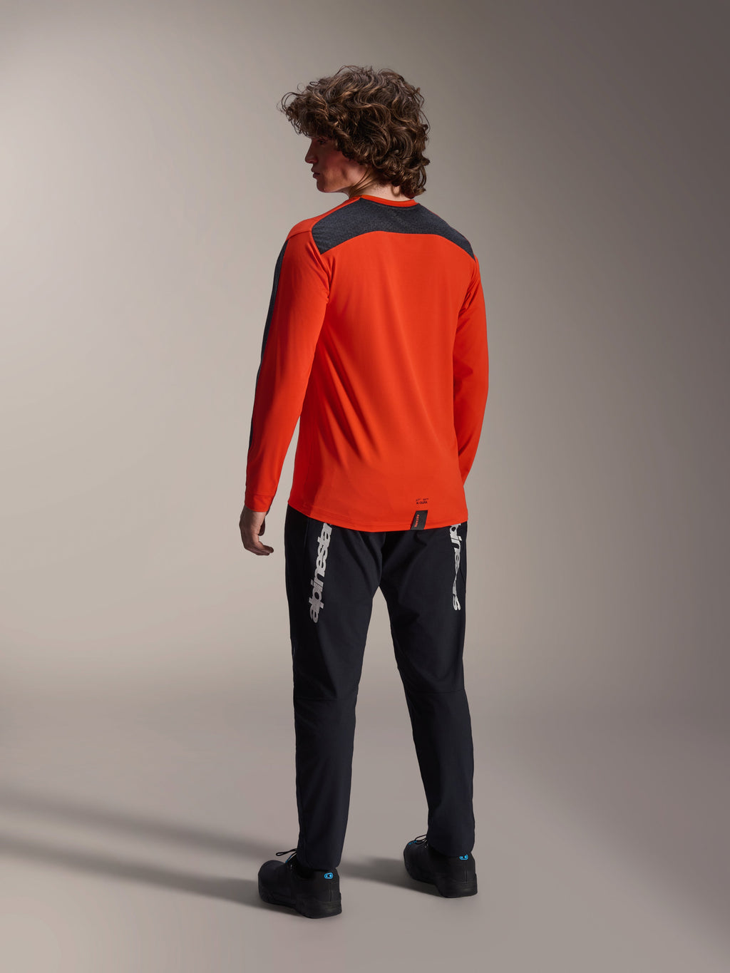 Vista traseira de um modelo a usar a camisola Alpinestars A-Dura Insert Astar em Spicy Orange, camisola de BTT de manga comprida com painel cinzento escuro nos ombros, combinada com calças pretas da marca, equipamento de ciclismo todo-o-terreno