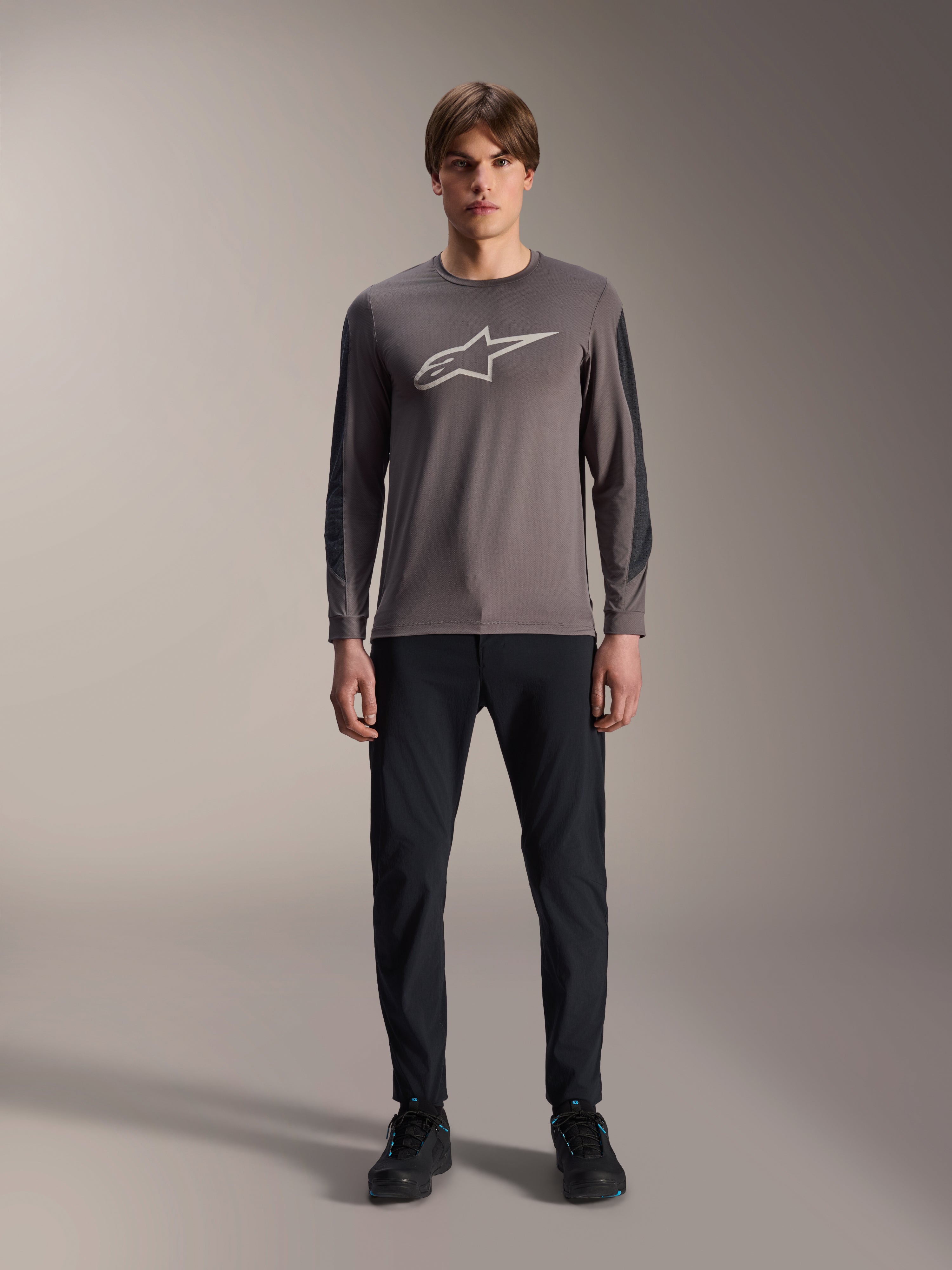 Camisola Alpinestars A-Dura Insert Astar, camisola de MTB de manga comprida, cinzento escuro, com um logótipo Astar cinzento claro proeminente no peito e inserções de manga texturadas mais escuras, vestida por um modelo com calças e sapatos pretos, equipamento de performance para BTT