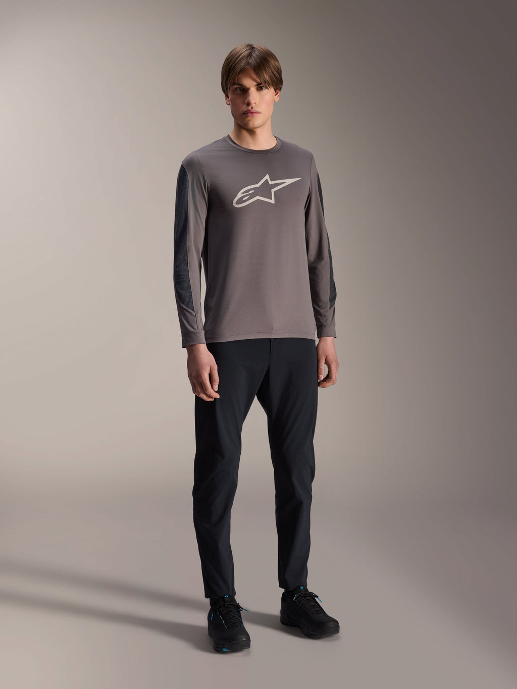 Alpinestars Camisola A-Dura Insert Astar, camisola de BTT de manga comprida, cinzento escuro, com grande logótipo branco no peito, usada com calças técnicas pretas e sapatos pretos, vestuário de ciclismo todo-o-terreno