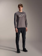 Alpinestars Camisola A-Dura Insert Astar, camisola de BTT de manga comprida, cinzento escuro, com grande logótipo branco no peito, usada com calças técnicas pretas e sapatos pretos, vestuário de ciclismo todo-o-terreno