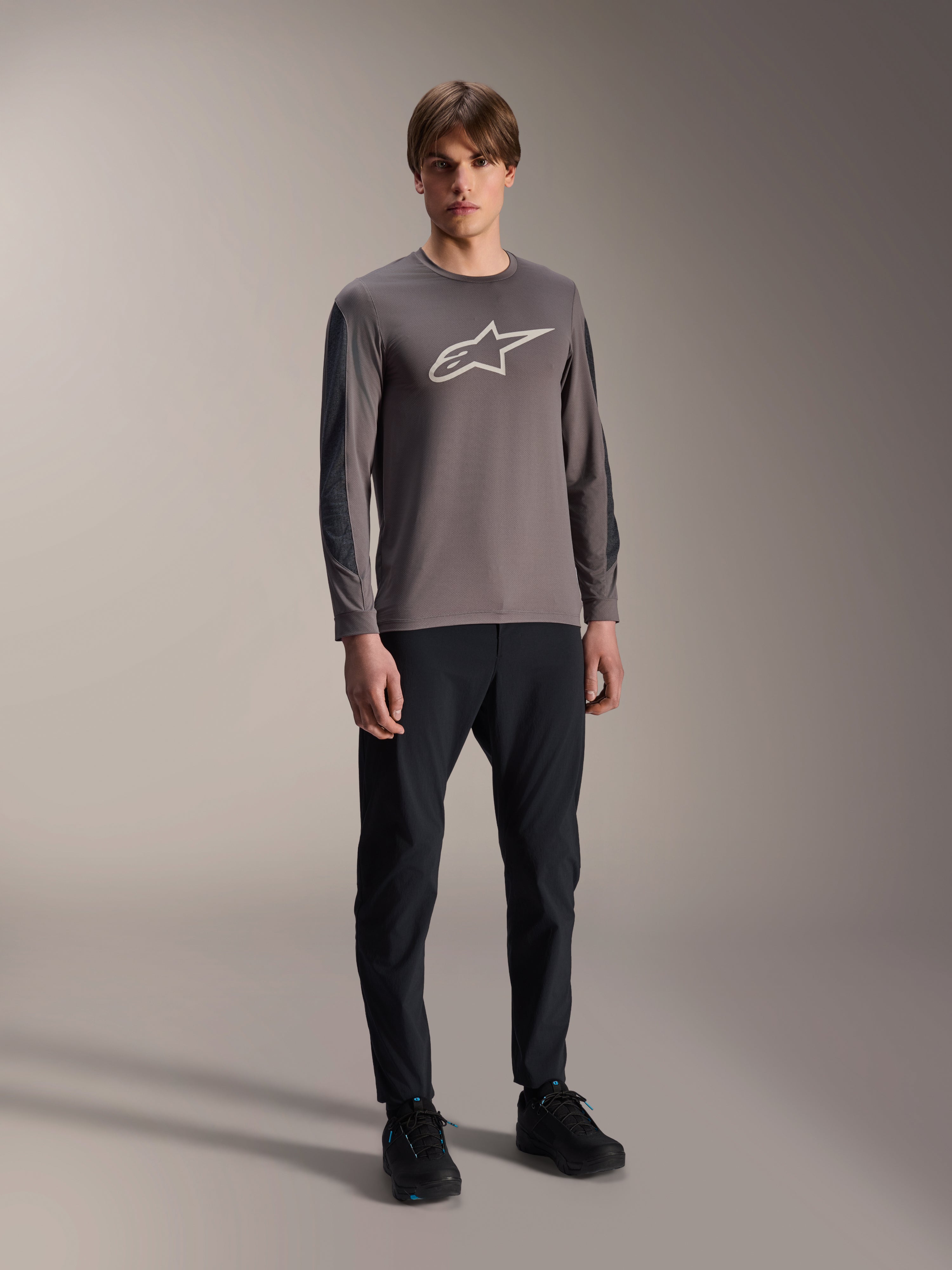 Alpinestars Camisola A-Dura Insert Astar, camisola de BTT de manga comprida, cinzento escuro, com grande logótipo branco no peito, usada com calças técnicas pretas e sapatos pretos, vestuário de ciclismo todo-o-terreno