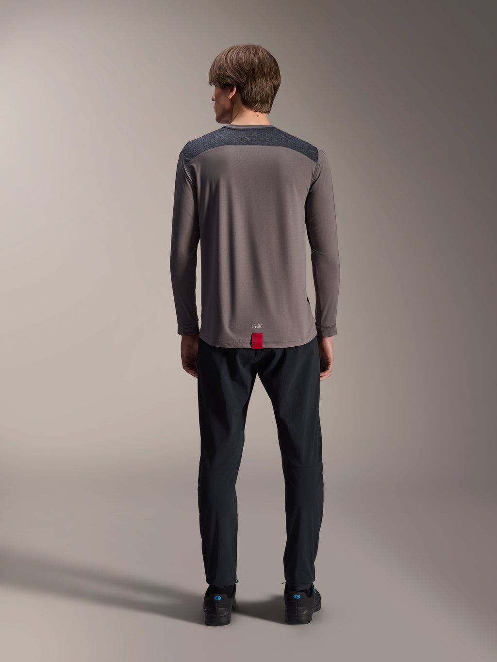 Camisola Alpinestars A-Dura Insert Astar de Manga Comprida em cinzento escuro, vista traseira, com painéis nos ombros em cinzento carvão e etiqueta vermelha na bainha, usada com calças e sapatos pretos, vestuário de performance para mountain bike
