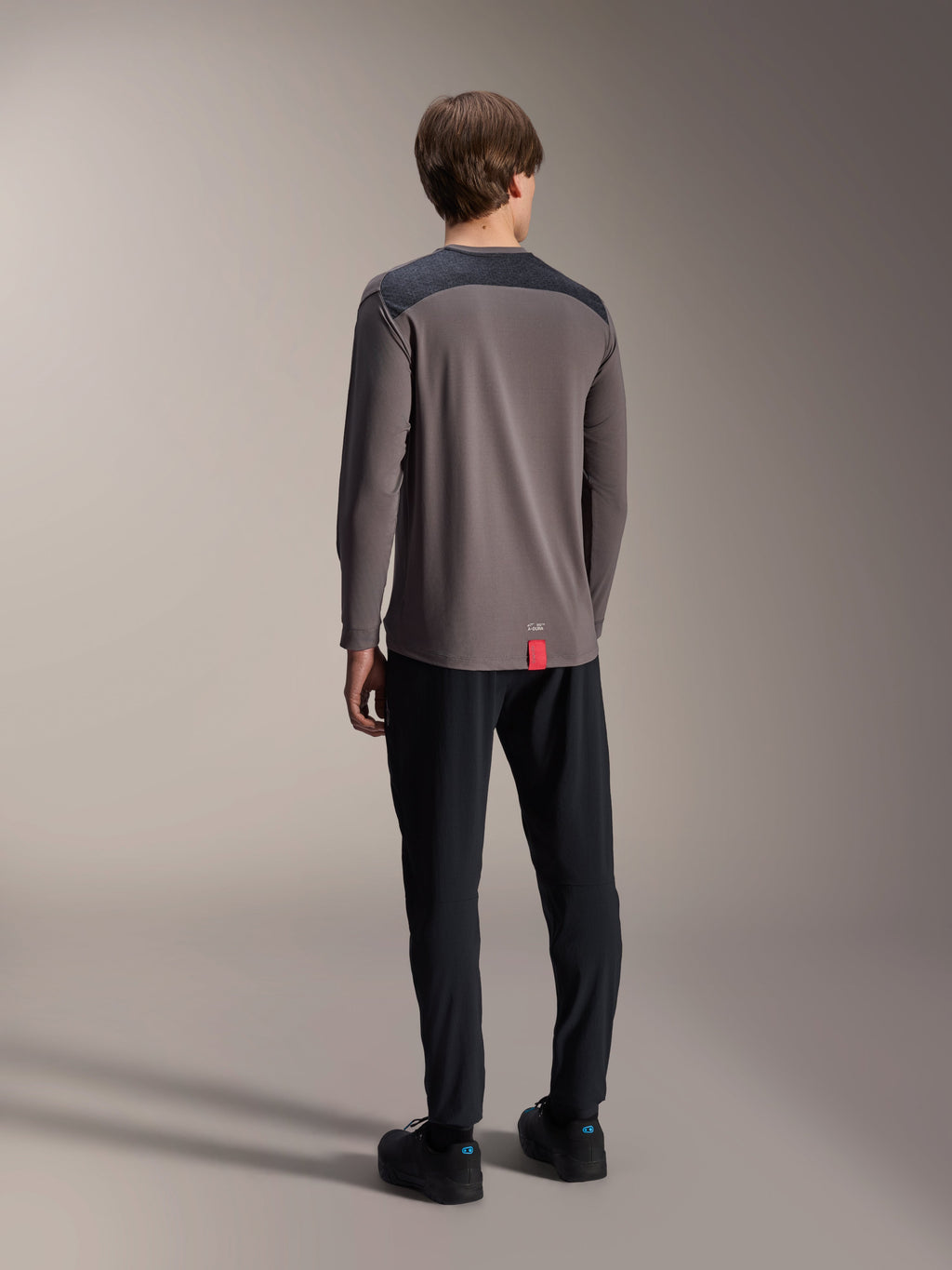Camisola Alpinestars A-Dura Insert Astar de Manga Comprida, Cinzento Escuro, vista traseira no modelo, com painéis de reforço nos ombros em cinzento carvão e uma pequena etiqueta vermelha na bainha, combinada com calças e sapatos pretos, vestuário técnico de MTB
