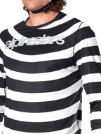 Primeiro plano da Camisola de Manga Comprida Alpinestars A-Aria Oscar em preto, apresentando riscas brancas horizontais com um gráfico espiral Oscar e um logótipo Alpinestars prateado refletor no peito, usada por um ciclista com a correia do capacete visível.