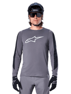 Alpinestars A-Dura Dri Astar Long Sleeve Jersey, Dark Gray, featuring a large white Astar logo on the chest and black mesh arm panels, worn by a rider with a black mountain bike helmet and performance sunglasses, MTB cycling apparel