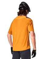 Camisola Alpinestars A-Aria Polartec Switch, camisola de MTB de manga curta, Dark Gold, vista traseira que mostra o ajuste de desempenho, usada com capacete preto e calções cinzentos, vestuário de trilhos durável e respirável