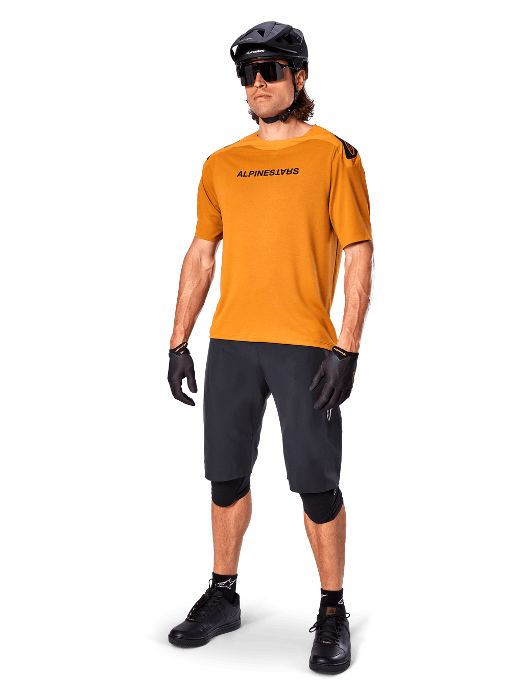 Ciclista masculino a usar a camisola Alpinestars A-Aria Polartec Switch em Dourado Escuro, camisola de BTT de manga curta, acompanhada por calções de ciclismo pretos, joelheiras, capacete preto e luvas, equipamento profissional de BTT para trilhos