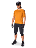Ciclista masculino a usar a camisola Alpinestars A-Aria Polartec Switch em Dourado Escuro, camisola de BTT de manga curta, acompanhada por calções de ciclismo pretos, joelheiras, capacete preto e luvas, equipamento profissional de BTT para trilhos