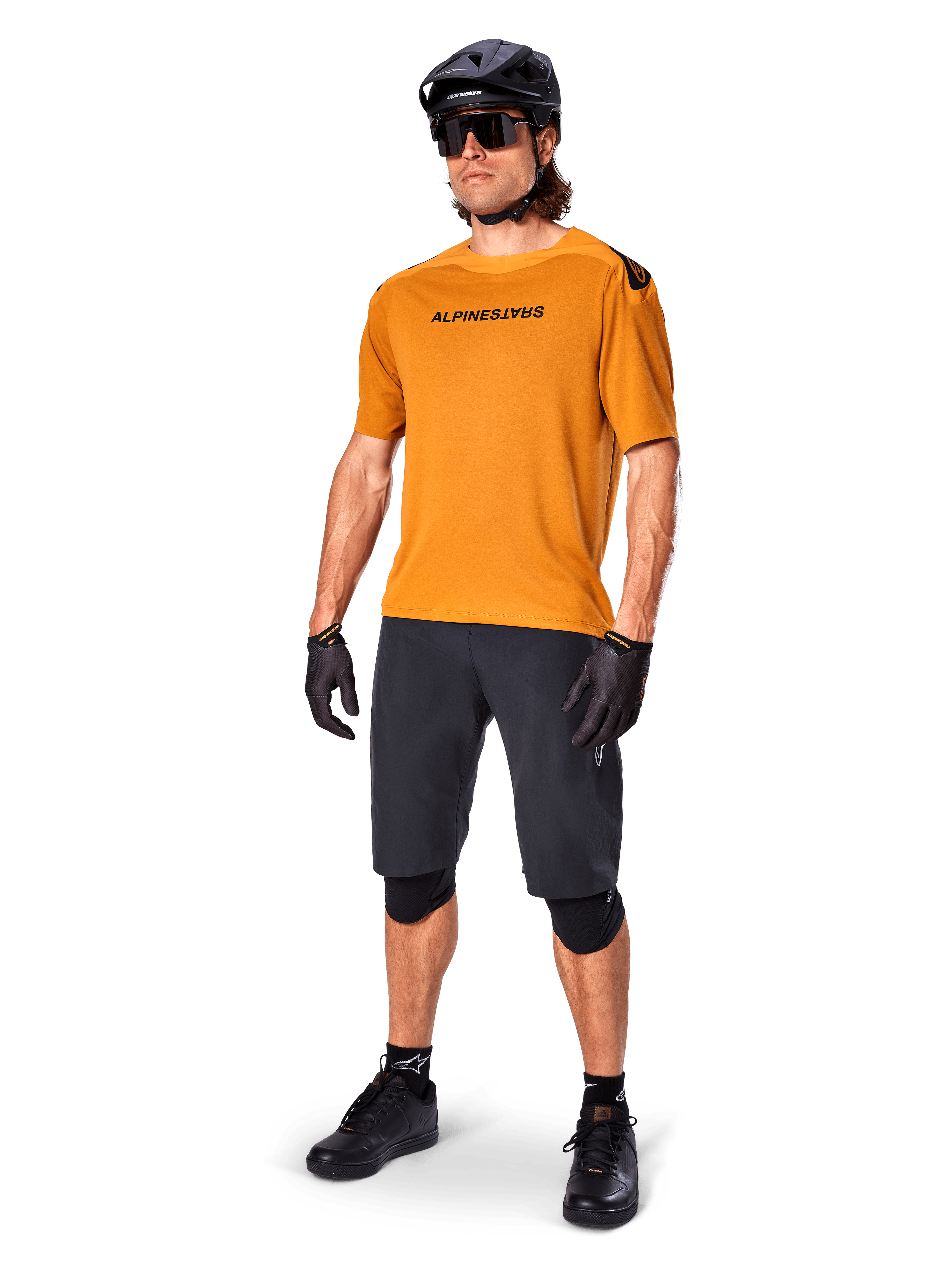 Ciclista masculino a usar a camisola Alpinestars A-Aria Polartec Switch em Dourado Escuro, camisola de BTT de manga curta, acompanhada por calções de ciclismo pretos, joelheiras, capacete preto e luvas, equipamento profissional de BTT para trilhos