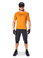 Ciclista masculino a usar a camisola de manga curta Alpinestars A-Aria Polartec Switch em Dark Gold, combinada com calções de BTT pretos, capacete, óculos de sol e luvas, equipamento profissional para trilhos de BTT