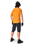 Alpinestars A-Aria Polartec Switch Jersey, camisola de BTT de manga curta, amarela, vista traseira de modelo a usar calções de BTT cinzento escuro, capacete preto e luvas, vestuário técnico de ciclismo para trilhos fora de estrada