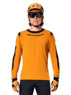 Modelo masculino a usar a camisola de manga comprida Alpinestars A-Dura Dri Wool em Dark Gold, coordenada com um capacete de BTT e luvas em dourado a condizer, painéis de malha pretos nos ombros e mangas, logótipo Alpinestars preto no peito, óculos de sol e calças escuras, vestuário técnico de BTT