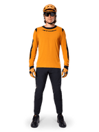 Ciclista de BTT com a Camisola de Manga Comprida Alpinestars A-Dura Dri Wool, Dark Gold com detalhes pretos no colarinho e nas mangas, combinada com calças pretas, capacete e luvas douradas a condizer e óculos de sol pretos, equipamento profissional de BTT