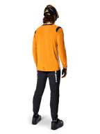 Ciclista de montanha visto de costas a usar a Camisola Alpinestars A-Dura Dri Wool de Manga Comprida em Dark Gold, calças técnicas pretas com logótipo branco e um capacete coordenado em preto e laranja, vestuário de BTT de alta performance.