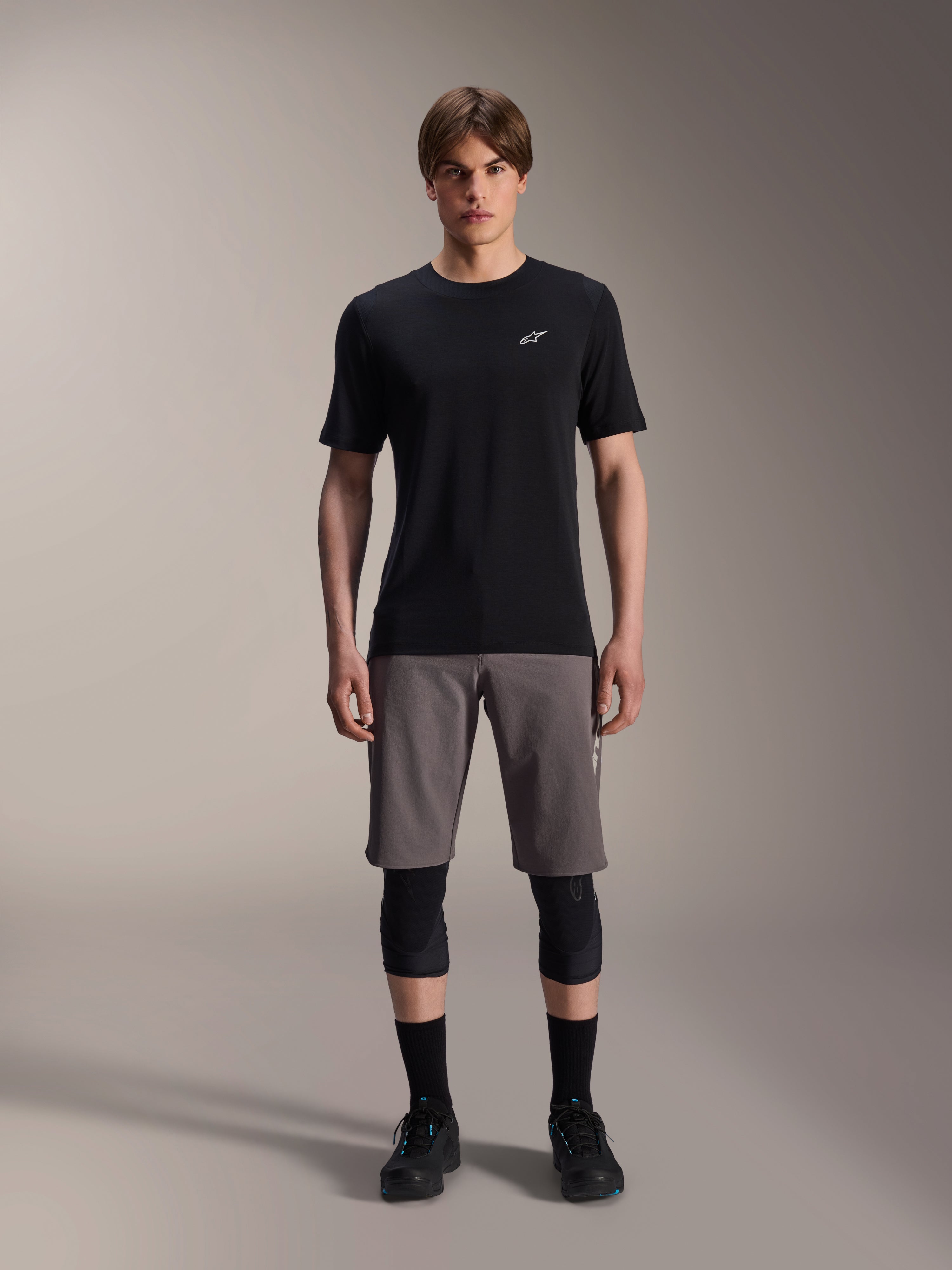 Modelo masculino a usar calções Alpinestars A-Dura Elite em cinzento escuro e camisola de manga curta A-Dura Dri-Wool em preto, combinados com joelheiras pretas e sapatos de ciclismo, vestuário técnico de mountain bike para performance fora de estrada.