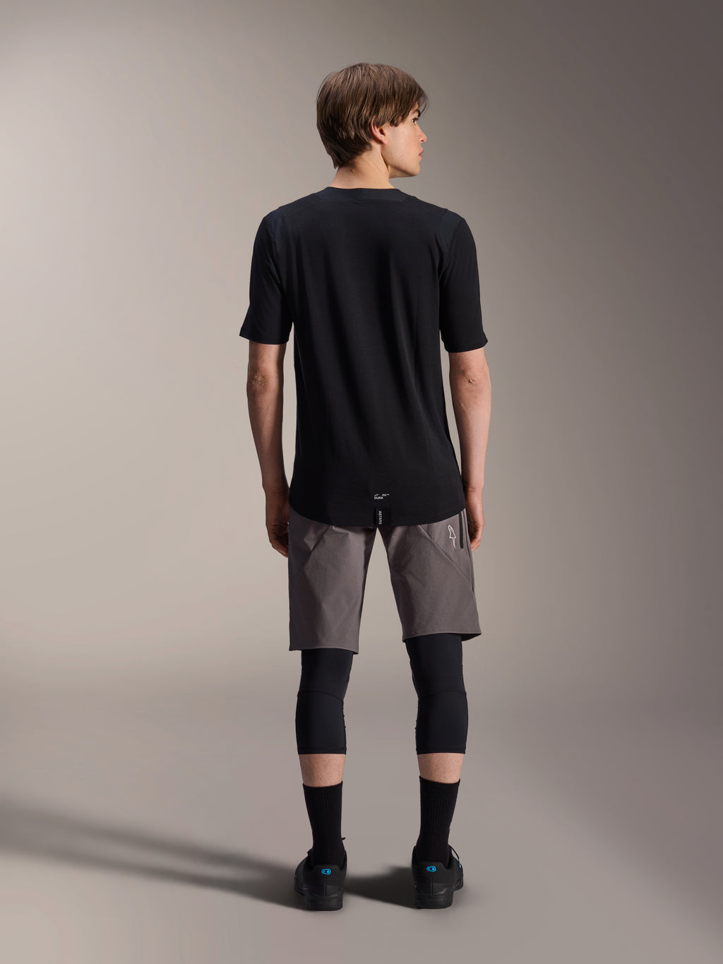 Calções Alpinestars A-Dura Elite, calções de mountain bike cinzento-escuros, vista traseira, usados com uma camisola preta de manga curta, joelheiras e meias pretas, equipamento técnico de ciclismo para condução off-road