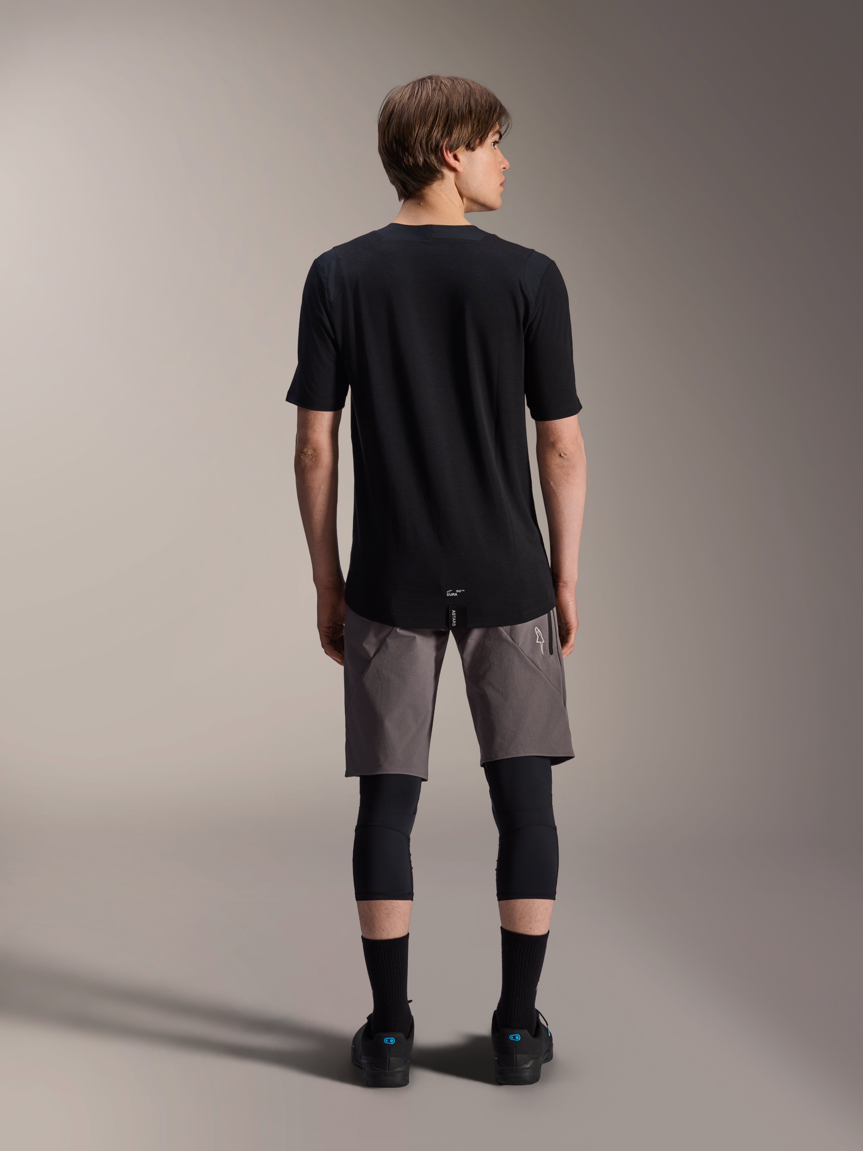 Calções Alpinestars A-Dura Elite, calções de mountain bike cinzento-escuros, vista traseira, usados com uma camisola preta de manga curta, joelheiras e meias pretas, equipamento técnico de ciclismo para condução off-road