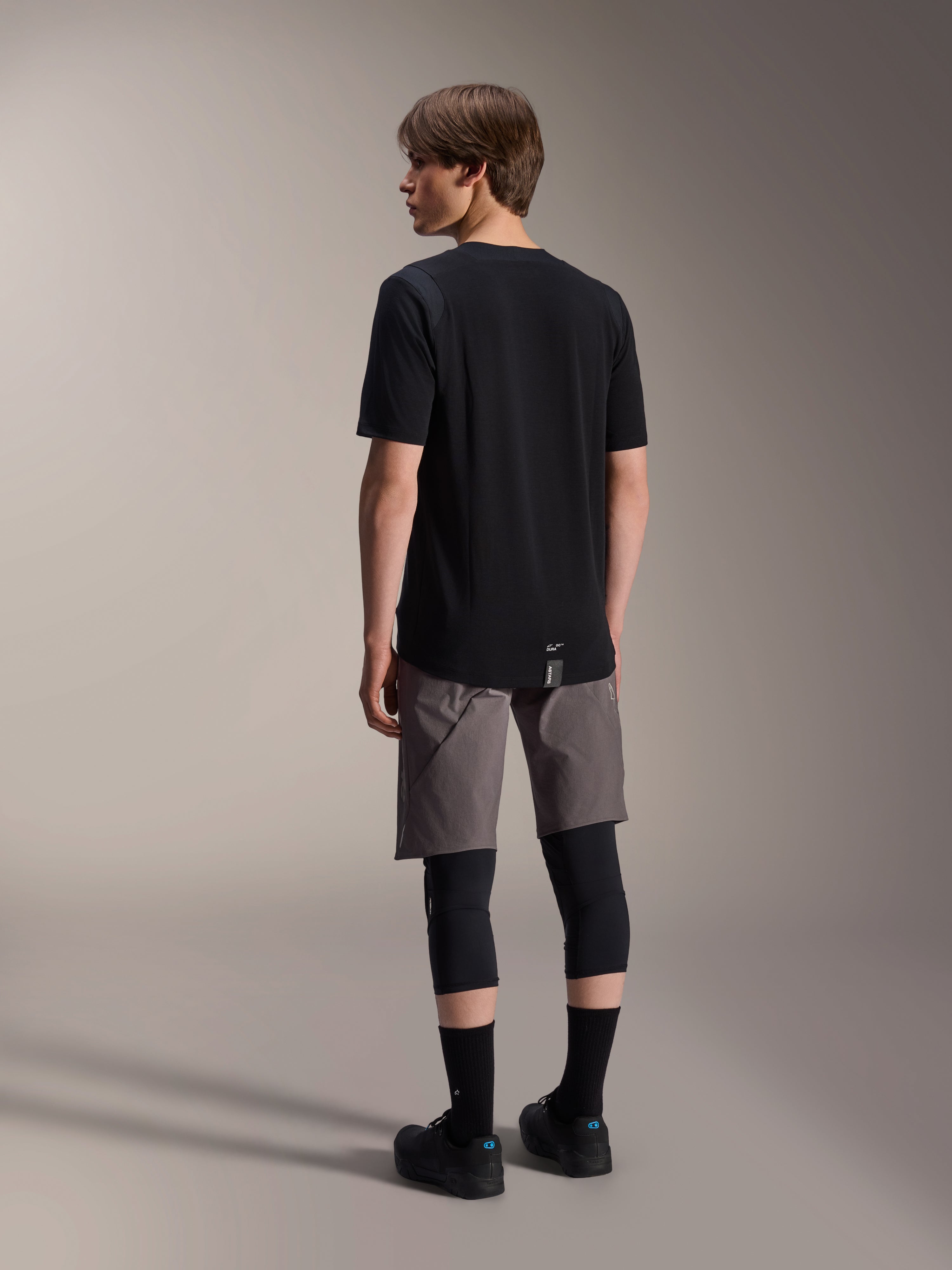 Camisola de Manga Curta Alpinestars A-Dura Dri Wool, preto, vista traseira no modelo, usada com calções de BTT cinzentos e joelheiras pretas, com bainha traseira alongada e logótipo subtil, vestuário técnico de BTT