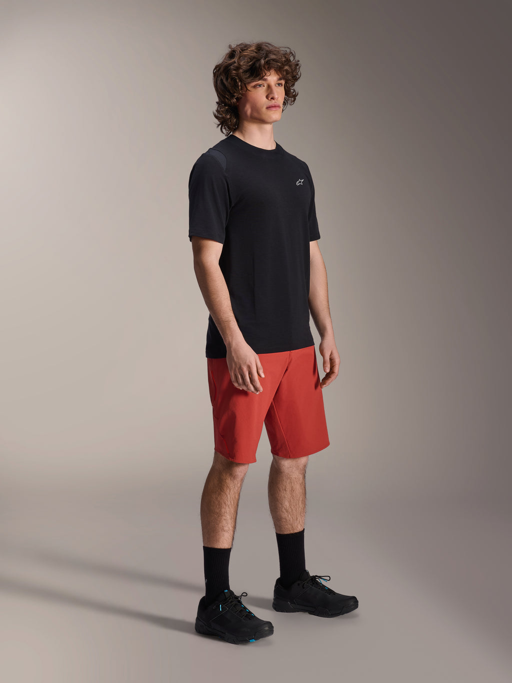 Modelo masculino a usar calções Alpinestars A-Dura Elite em Laranja Escuro e jersey de manga curta A-Dura Dri Wool em preto, equipamento de performance para BTT, combinado com meias e sapatos pretos, design atlético para ciclismo fora de estrada.