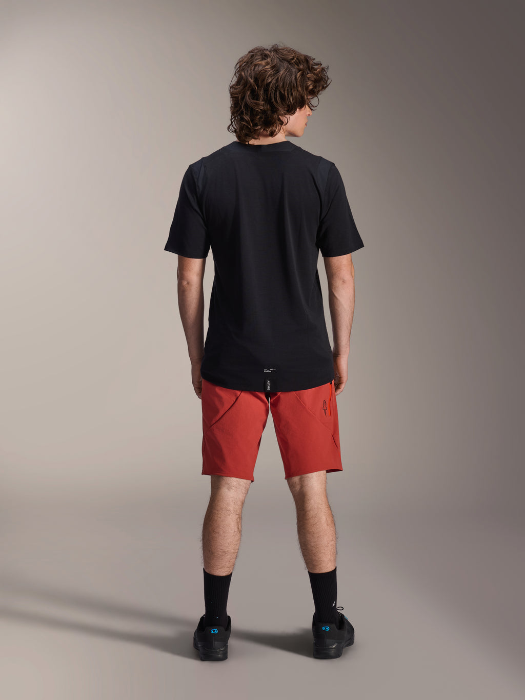 Modelo masculino a usar calções Alpinestars A-Dura Elite em Dark Orange e camisola de manga curta A-Dura Dri Wool em preto, visto de costas, combinado com meias pretas e sapatos de ciclismo, vestuário profissional de mountain bike