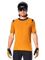 Camisola de Manga Curta Alpinestars A-Dura Dri Wool, dourado escuro, usada com capacete de MTB preto, óculos de sol e luvas, com gola e painéis nos ombros pretos, mistura de lã drirelease respirável para mountain biking