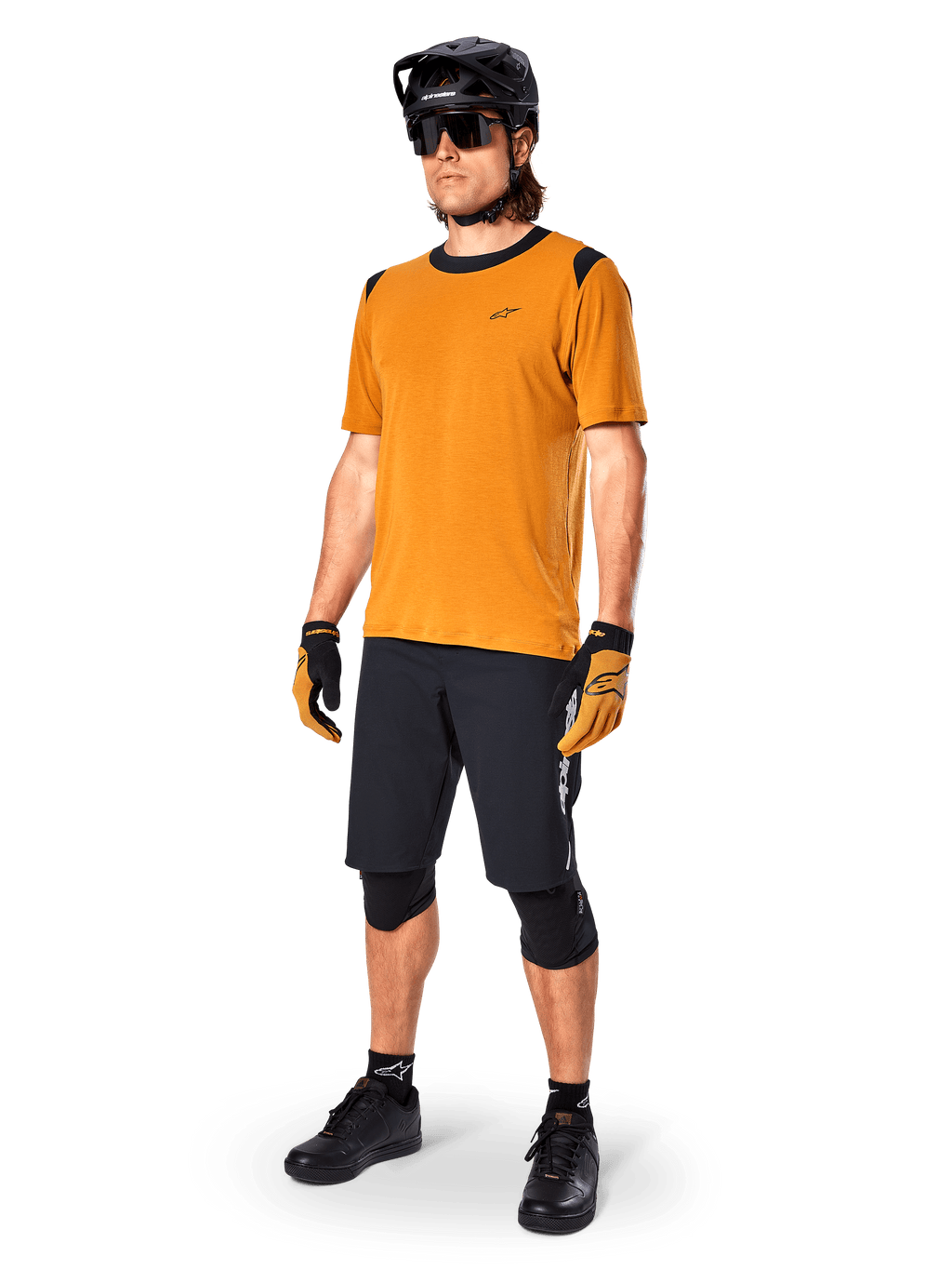 Modelo masculino a usar a camisola de manga curta Alpinestars A-Dura Dri Wool em dourado escuro, camisola de BTT com detalhes pretos nos ombros e gola, combinada com calções de BTT pretos, capacete preto e óculos de sol, equipamento técnico de ciclismo fora de estrada