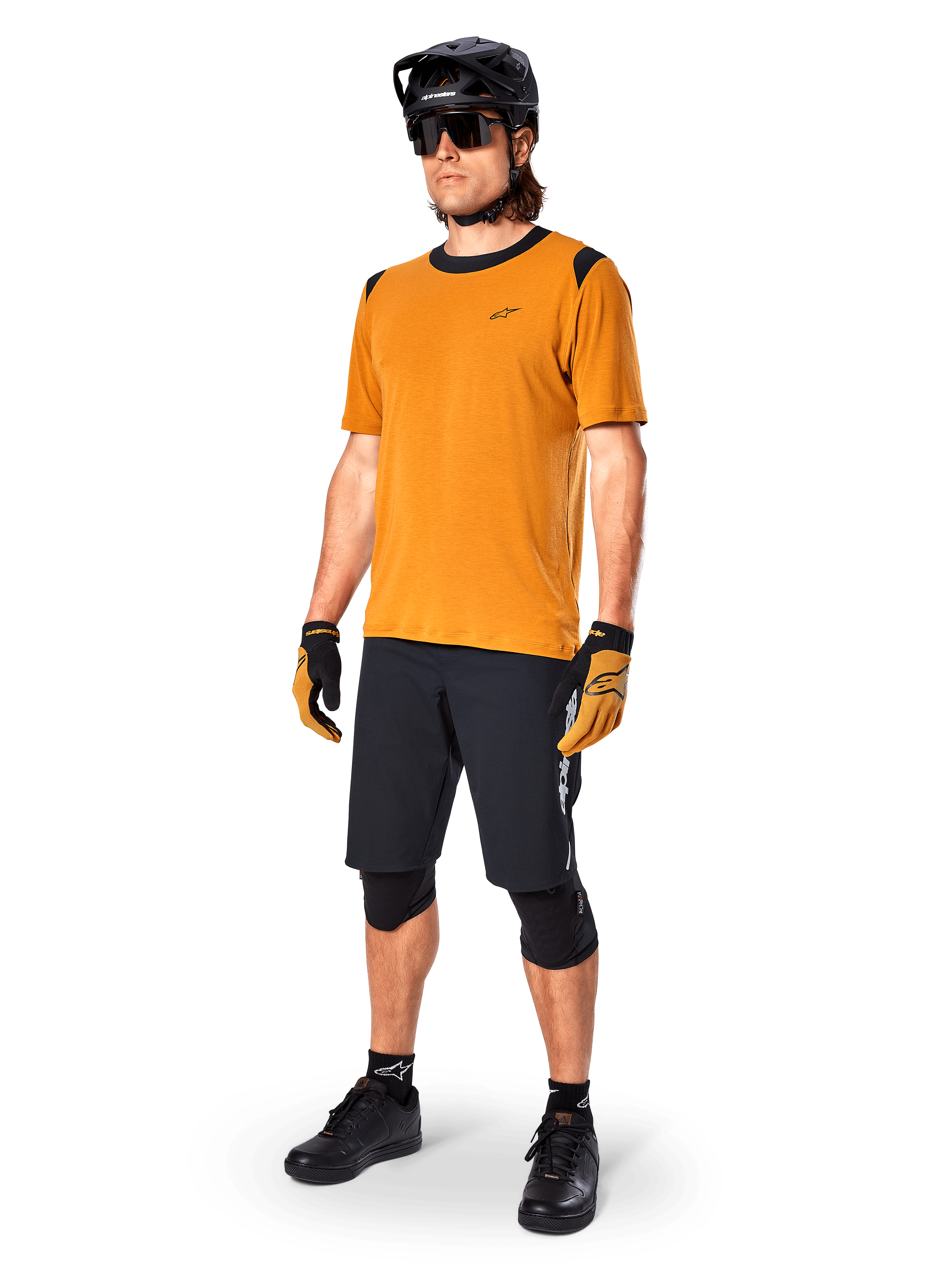 Modelo masculino a usar a camisola de manga curta Alpinestars A-Dura Dri Wool em dourado escuro, camisola de BTT com detalhes pretos nos ombros e gola, combinada com calções de BTT pretos, capacete preto e óculos de sol, equipamento técnico de ciclismo fora de estrada