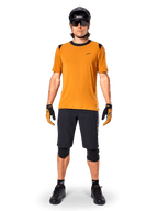 Camisola de Manga Curta Alpinestars A-Dura Dri Wool em Dark Gold, camisola de mountain bike usada com calções pretos e equipamento de proteção, tecido amarelo mostarda com gola e detalhes pretos nos ombros, vestuário de ciclismo de performance para trilhos.