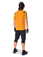 Camisola de Manga Curta Alpinestars A-Dura Dri Wool, camisola de ciclismo, Dark Gold, vista traseira de ciclista de montanha com camisola amarelo-mostarda e detalhes em malha preta nos ombros, combinada com calções pretos, joelheiras, capacete preto e luvas, equipamento técnico de ciclismo todo-o-terreno