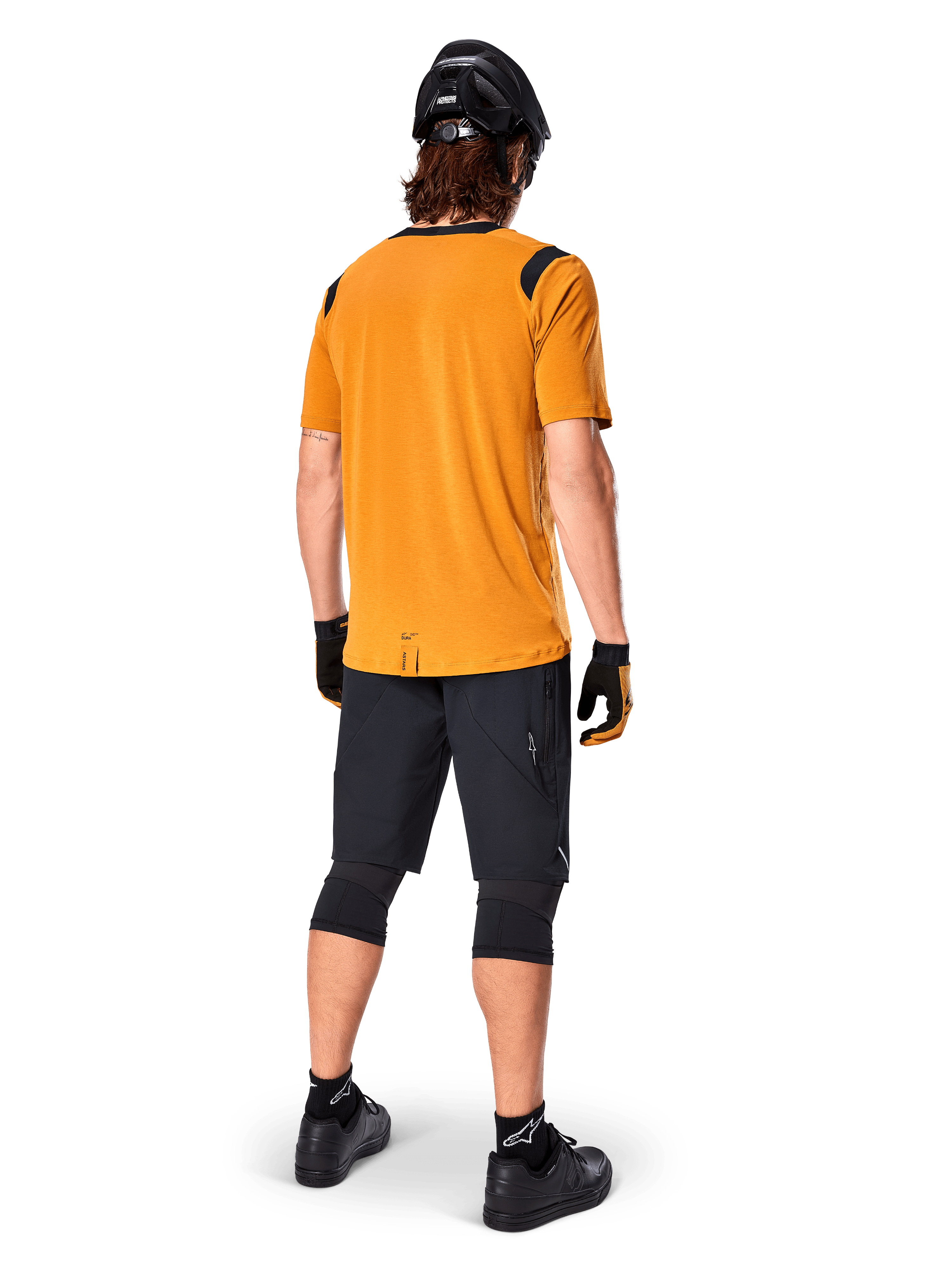 Camisola de Manga Curta Alpinestars A-Dura Dri Wool, camisola de ciclismo, Dark Gold, vista traseira de ciclista de montanha com camisola amarelo-mostarda e detalhes em malha preta nos ombros, combinada com calções pretos, joelheiras, capacete preto e luvas, equipamento técnico de ciclismo todo-o-terreno