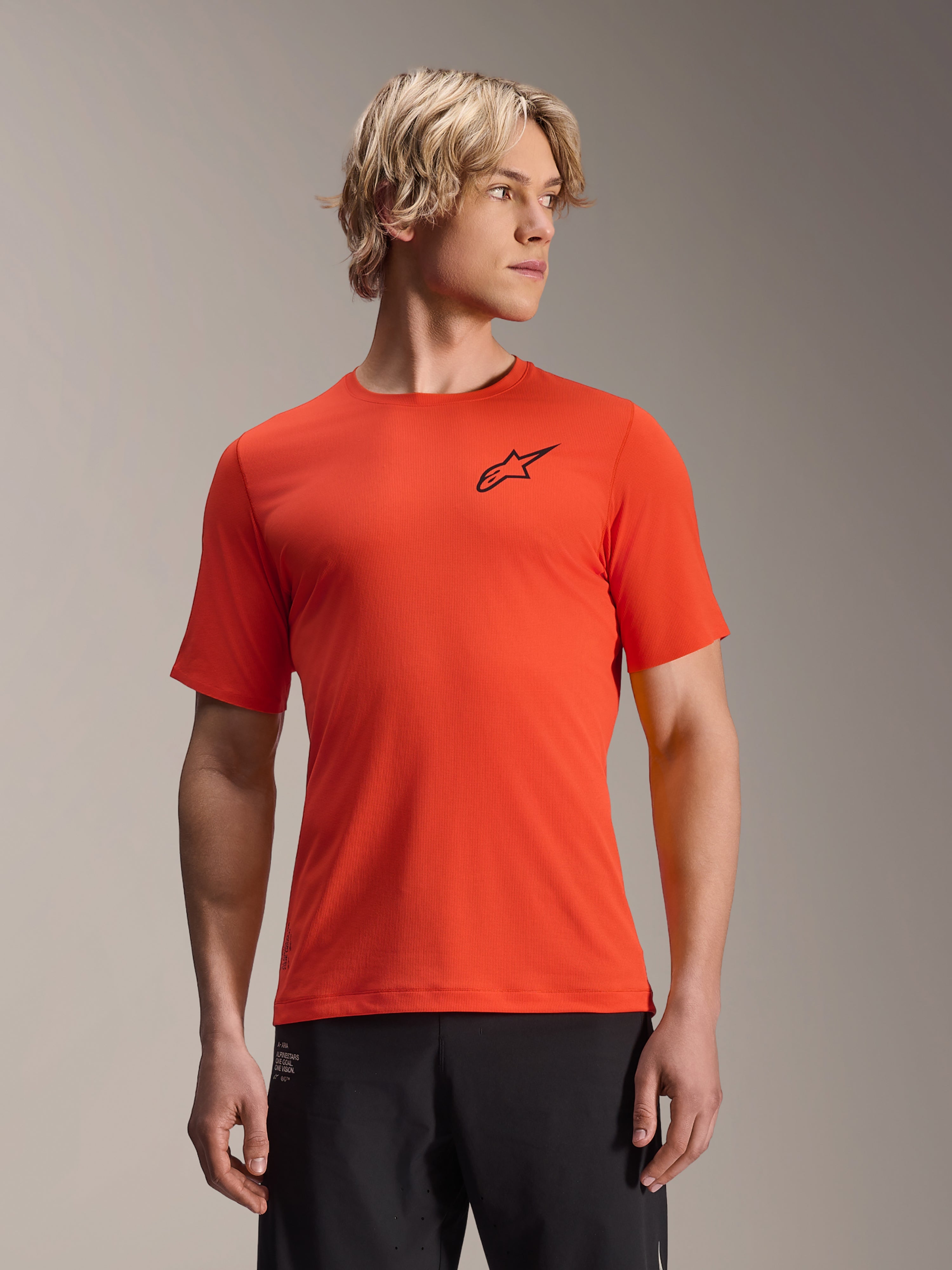 Alpinestars A-Aria Astar Jersey de Manga Curta, Spicy Orange com logótipo de estrela preto, camisola de BTT para homem, corte de performance, vestida por modelo masculino de cabelo loiro e calças pretas