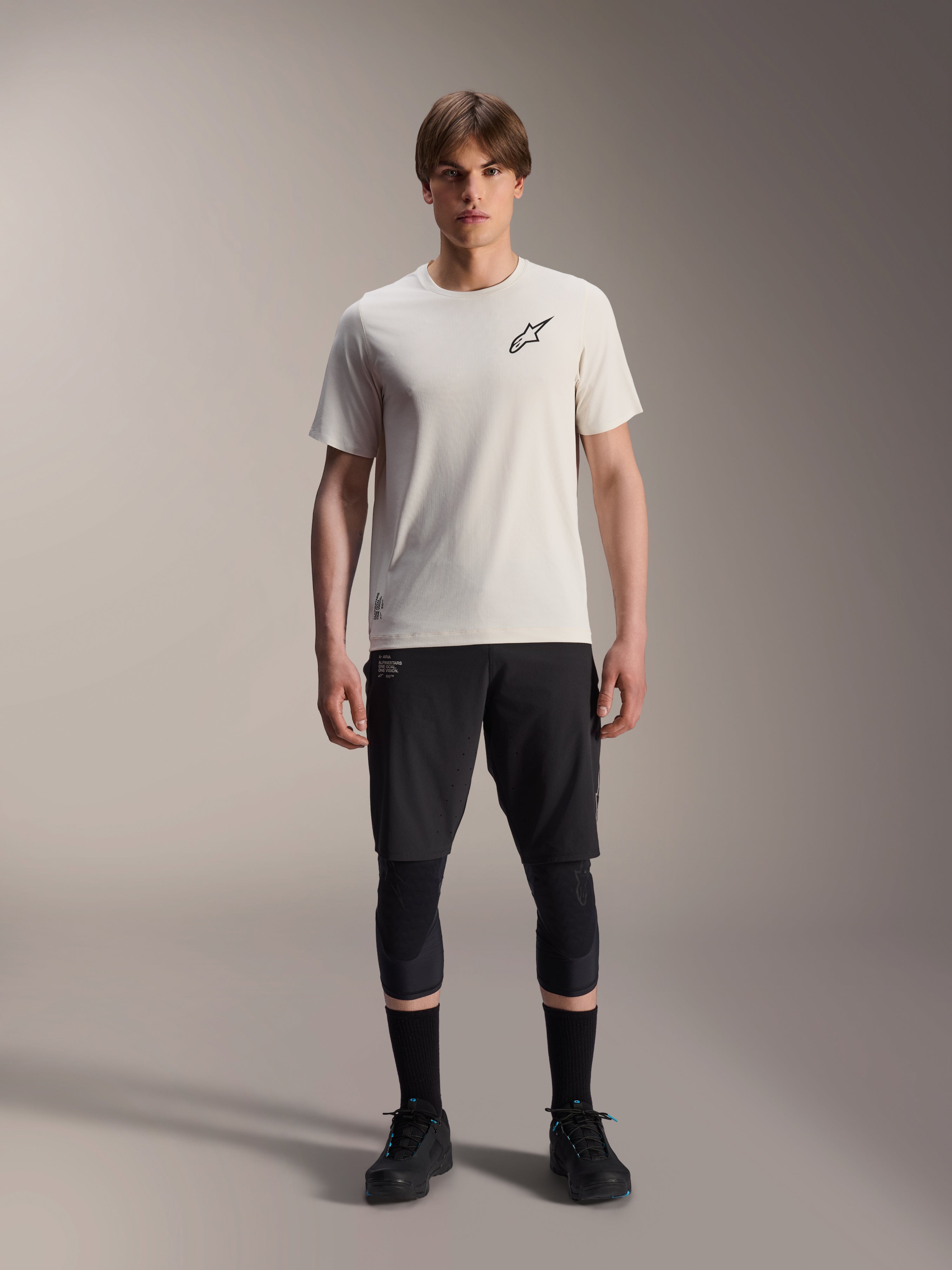 Modelo masculino a usar a Camisola de Manga Curta Alpinestars A-Aria Astar em Ecru, combinada com calções de BTT pretos, joelheiras e sapatos de ciclismo pretos, apresentando um logótipo de estrela preto minimalista no peito, concebida para trilhos todo-o-terreno