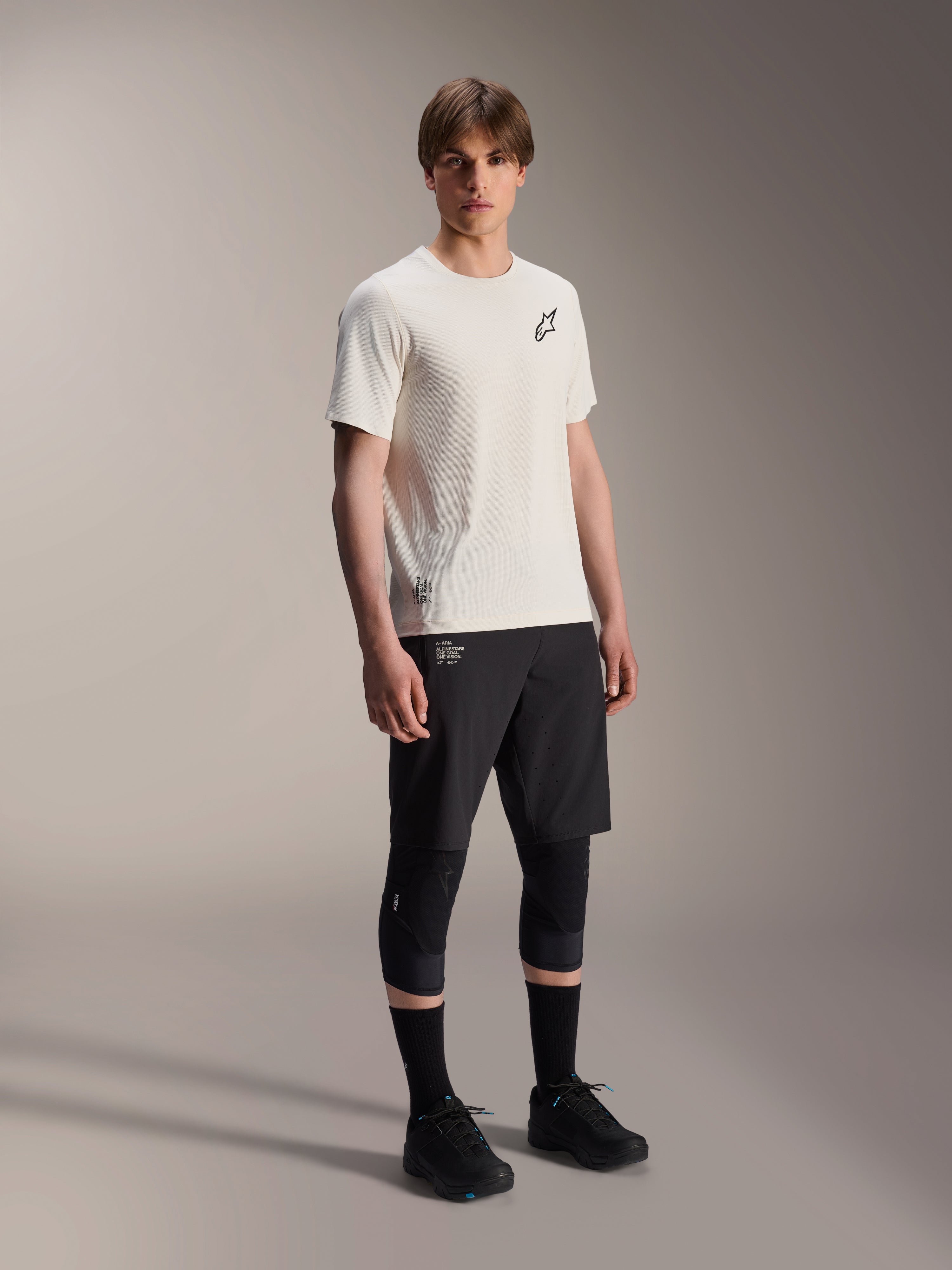 Camisola Alpinestars A-Aria Astar, Manga Curta, Ecru, com logótipo preto no peito, usada com calções de ciclismo pretos e joelheiras, equipamento de performance leve para MTB