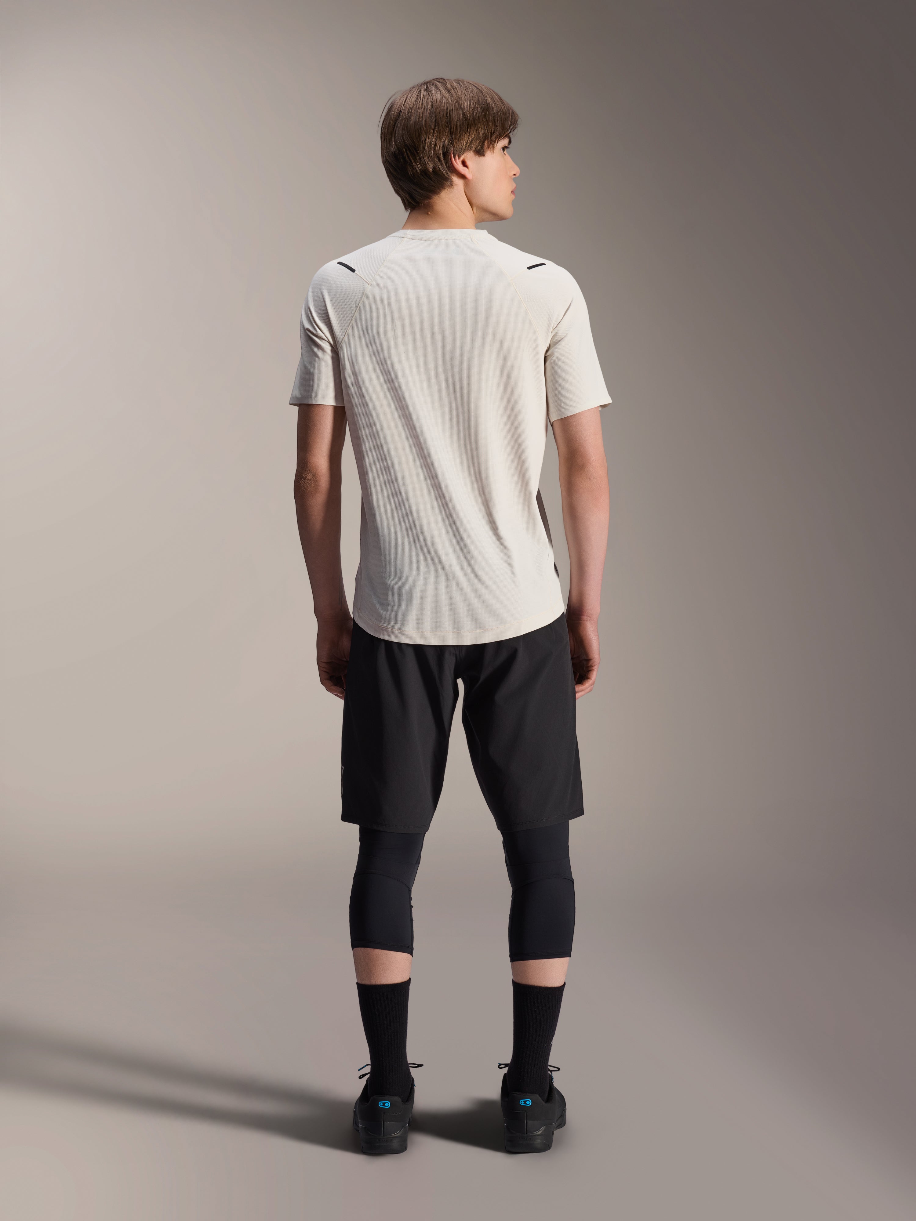 Modelo masculino de costas a usar a Camisola de Manga Curta Alpinestars A-Aria Astar em Ecru, combinada com calções de BTT pretos e leggings de proteção, com cauda mais comprida e ajuste de performance para trilhos todo-o-terreno.