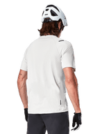 Alpinestars A-Aria Astar Jersey, Manga Curta, White Dove, vista traseira de camisola de BTT leve e respirável, usada com um capacete branco e luvas de ciclismo pretas, com cauda mais longa para maior cobertura