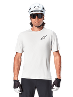 Ciclista a usar a camisola de manga curta Alpinestars A-Aria Astar, cor White Dove com um pequeno logótipo Astar preto no peito, capacete de ciclismo branco, óculos de sol pretos e luvas pretas e brancas, equipamento de performance para MTB e condução fora de estrada