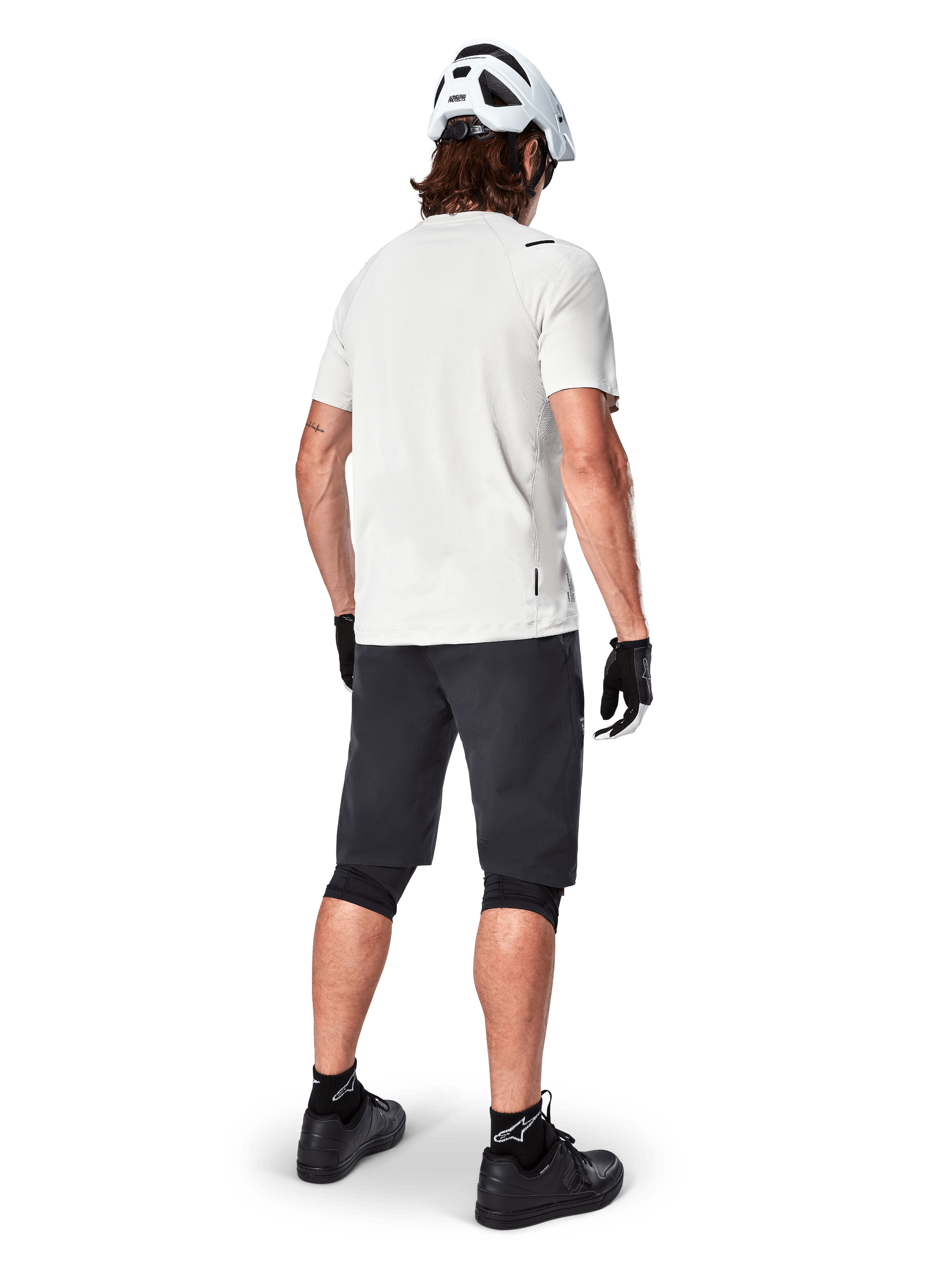 Ciclista a usar a camisola de manga curta Alpinestars A-Aria Astar em White Dove, vista traseira, combinada com calções de BTT pretos, joelheiras e um capacete branco, equipamento leve e respirável para BTT fora de estrada