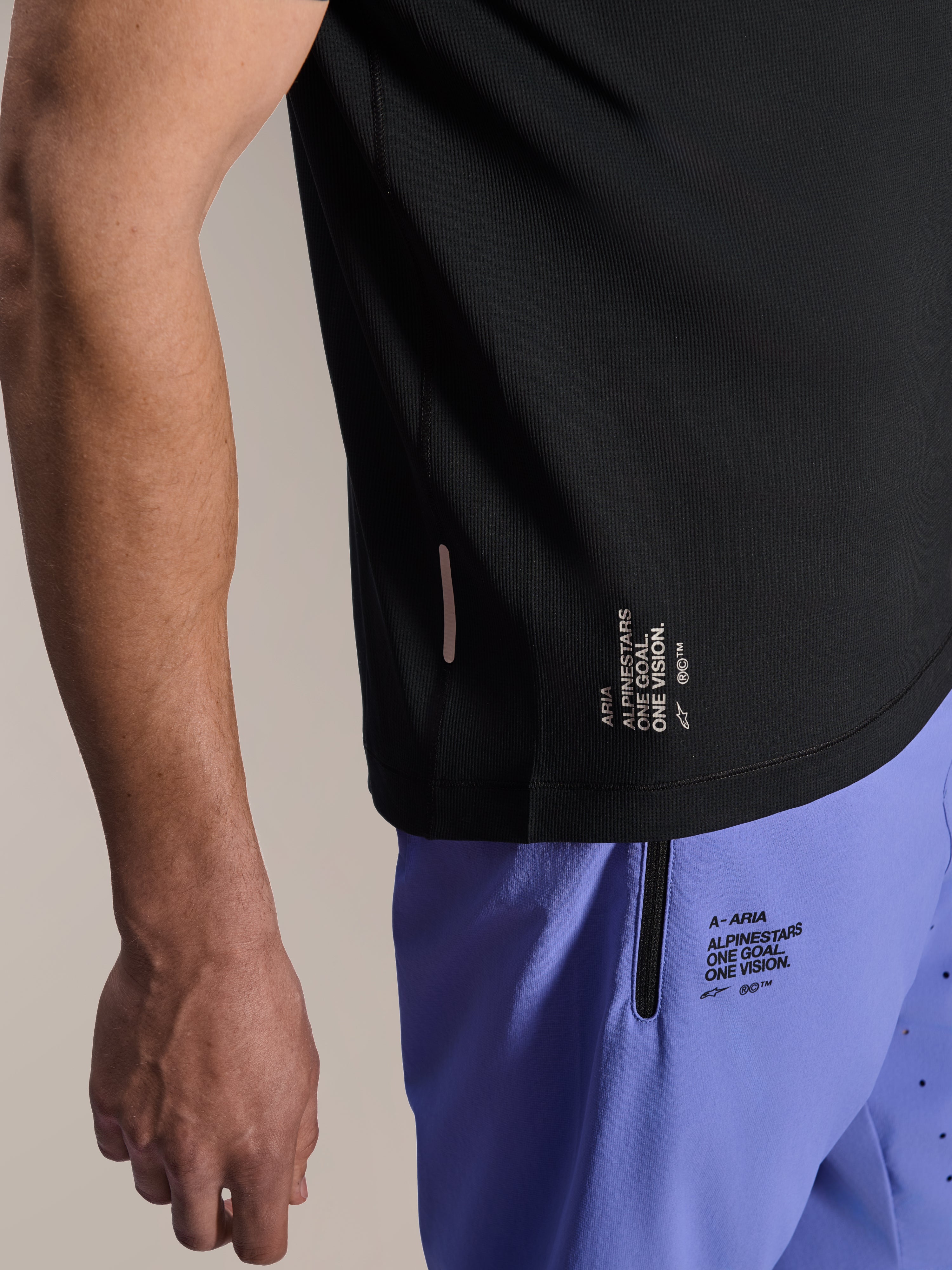 Camisola de Manga Curta Alpinestars A-Aria Switch, camisola de BTT preta, grande plano da bainha lateral com a marca branca 'ONE GOAL. ONE VISION.', usada com calções de ciclismo roxo claro com logótipos a condizer