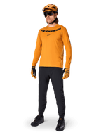 Camisola de Manga Comprida Alpinestars A-Dura Rocker, camisola de ciclismo em Dark Gold, usada com capacete de BTT a condizer e calças pretas, com logótipo da marca a preto no peito, equipamento de performance coordenado para ciclismo de montanha fora de estrada
