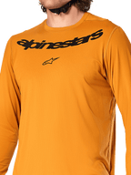 Camisola de manga comprida Alpinestars A-Dura Rocker, ouro escuro, vista detalhada do peito com marca nominativa e logótipo de estrela Alpinestars em preto, tecido jacquard texturizado com absorção de humidade, vestuário de performance para ciclismo MTB
