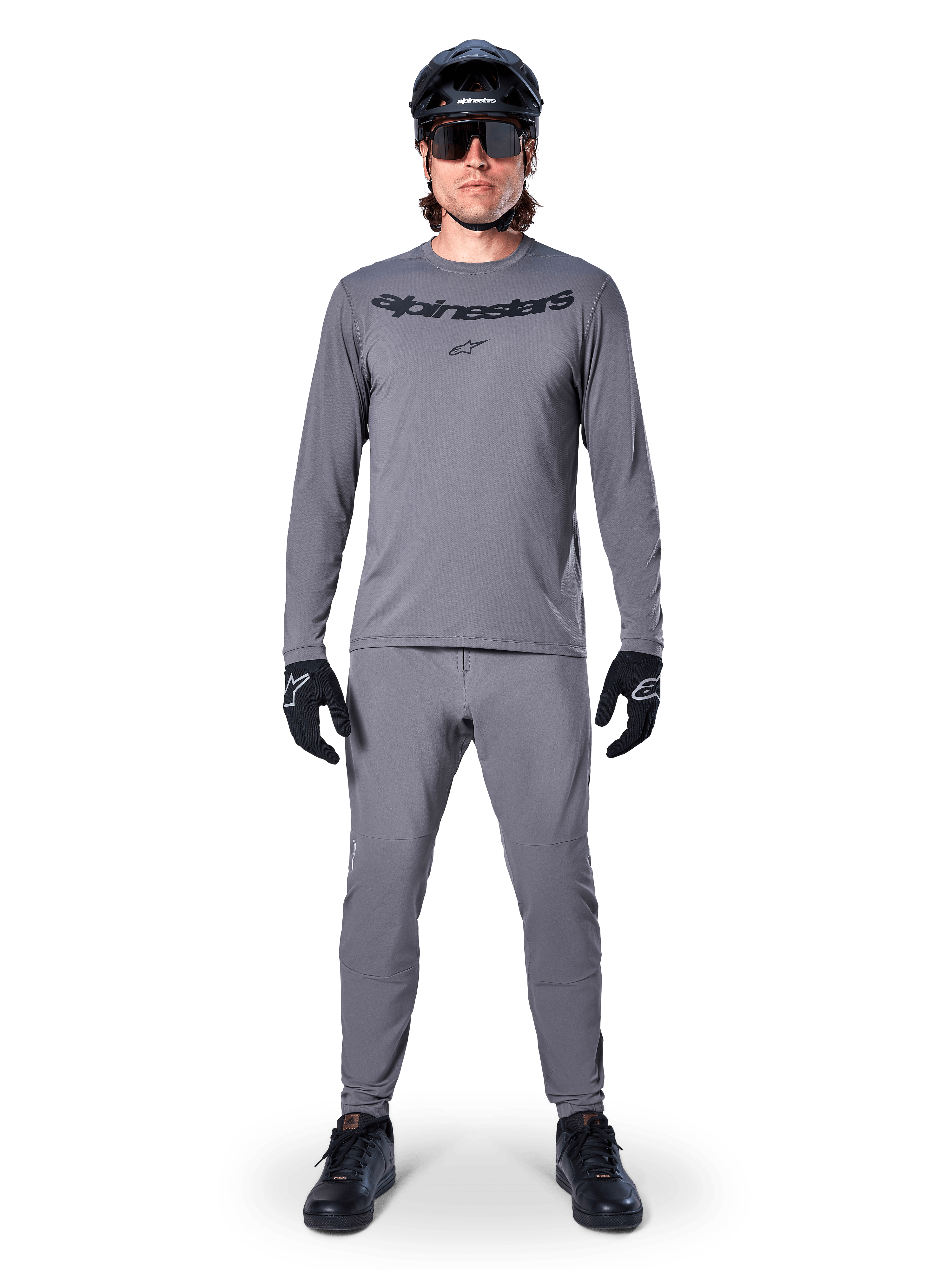 Male cyclist wearing Alpinestars A-Dura Rocker Long Sleeve Jersey in Dark Gray, matching gray pants, black MTB helmet, sunglasses and gloves, professional mountain bike apparel for off-road trails