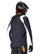 Ciclista de BTT a usar a Camisola de Manga Longa Alpinestars A-Supra Oscar, preta com painéis brancos nos ombros e mangas, vista de costas, capacete e luvas pretas, calças com detalhes laranja, equipamento de ciclismo de montanha durável